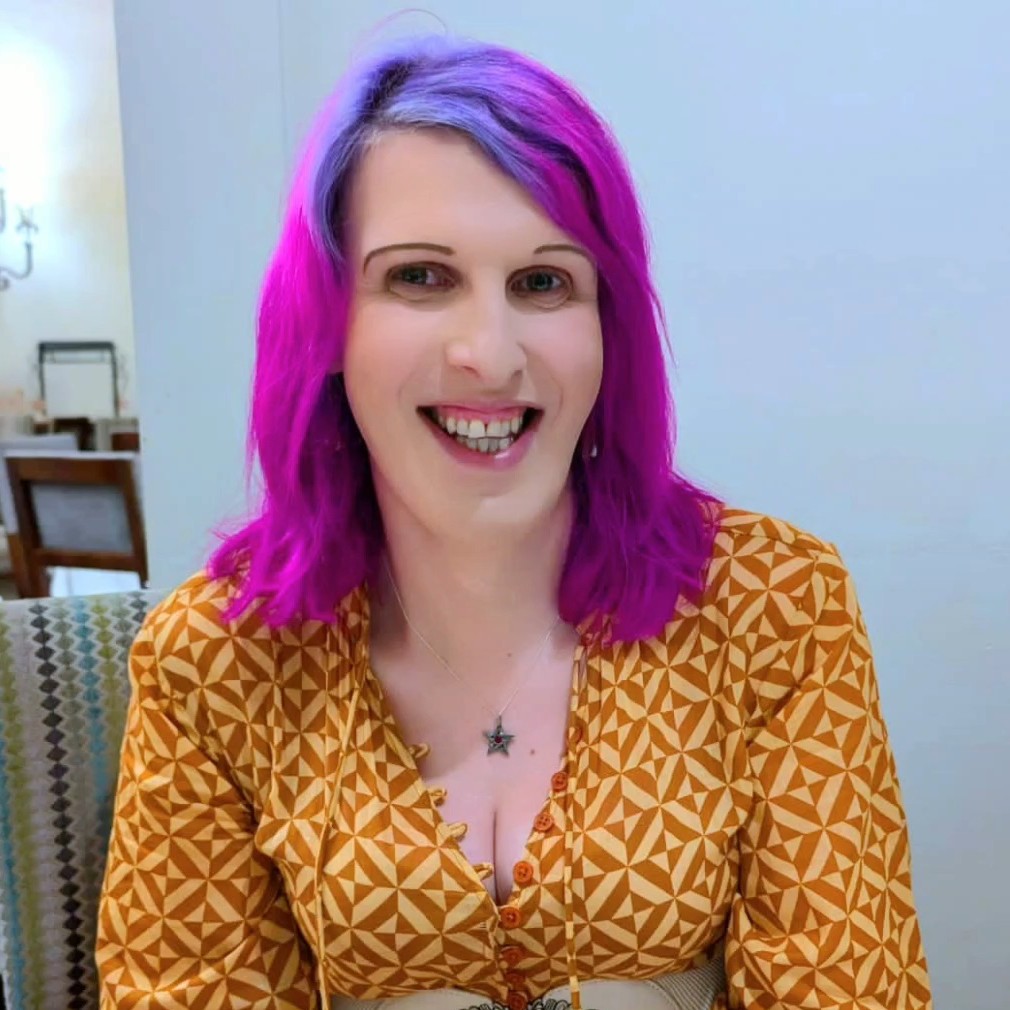 Head and Shoulders image of a smiling Tara Hewitt. Tara is wearing a blouse with a yellow ansd purple geometric design and she has shoulder-length purple coloured hair